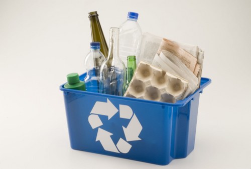 A blue plastic recycling bin filled with assorted waste items including transparent and green glass bottles, a clear plastic water bottle with a blue cap, an egg carton, a rolled newspaper, and other paper materials. The bottles are positioned upright and leaning against each other, with some partially obscured. The bin features a white recycling symbol on its front and is placed on a plain, light-colored surface with a neutral background. The environment appears to be an indoor space, possibly related to waste collection or rubbish removal services in Staines, with the focus on waste segregation and disposal. The image reflects typical contents handled by office clearance companies such as Office Clearance Staines, emphasizing the importance of proper rubbish disposal in local waste management practices.