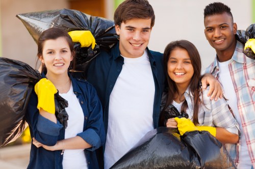A group of four young volunteers standing outdoors in a clean, well-lit environment, each holding large black rubbish bags filled with waste. The individuals, two women and two men, are smiling and dressed casually; some are wearing bright yellow gloves. They appear to have participated in a community or environmental cleanup activity, with the rubbish bags held over their shoulders and in front of them, ready for disposal. The background features a blurred outdoor setting with greenery and a pathway, indicative of a park or residential area, likely in the Staines region. The scene reflects a positive and collaborative effort toward waste removal and environmental responsibility, with a focus on rubbish collection and disposal, aligning with services offered by companies like Office Clearance Staines.