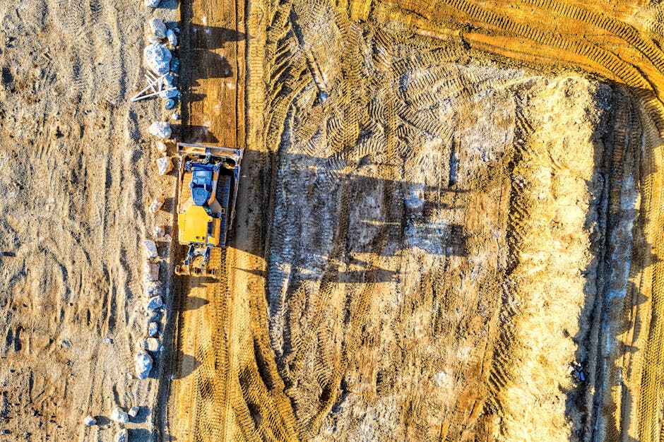 An aerial view of a construction site featuring a yellow excavator with a black arm, positioned in the center of the frame. The excavator is working on a large, partially excavated area with visible t