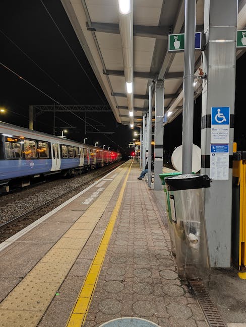 A railway station platform at night, illuminated by overhead lighting, showing a row of parked trains on the left side with blue and white exterior finishes. The platform has tactile paving in yellow