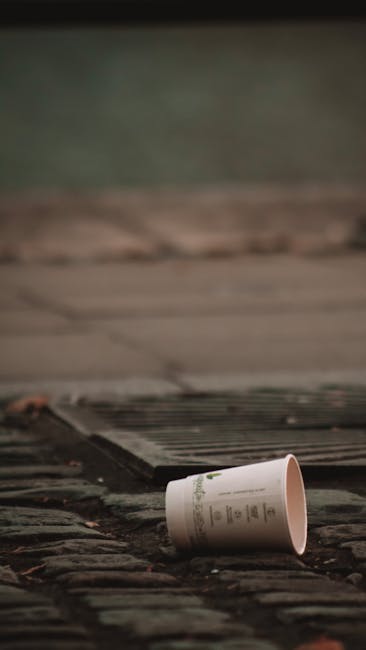 A photograph showing two wheeled rubbish bins positioned side by side on a cobblestone pavement against a plain, light-colored exterior wall. The bin on the left is dark green with a textured plastic